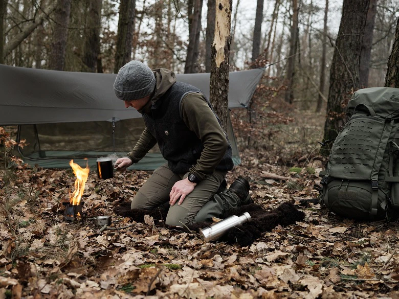 Person beim Bushcrafting an einem Lagerfeuer, im Hintergrund Tarp und Rucksack