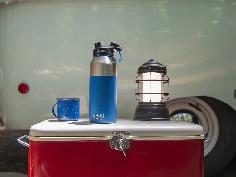 Outdoor Imagebild - blaue Camelbak Isolierflasche steht auf einer Campingbox neben einem blauen Becker und einer Outdoorlaterne. Im Hinergrund sind Umrisse eines Vans zu erkennen.