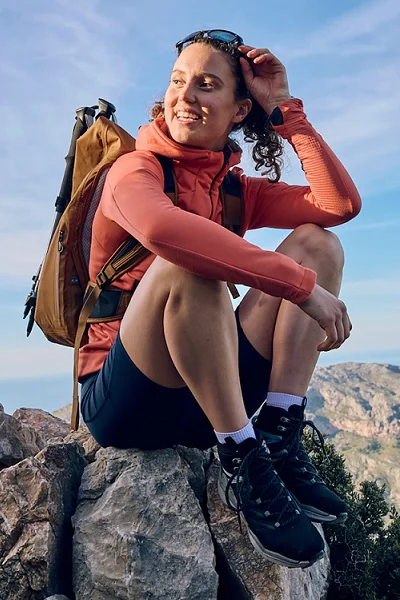 Outdoor Imagebild - Person sitzt im freien auf einem Felsen und trägt eine rote Jacke, blaue Shorts sowie einen Rucksack. Die Schuhe sind ein Paar Lowa Maddox Pro Mid GTX in blau