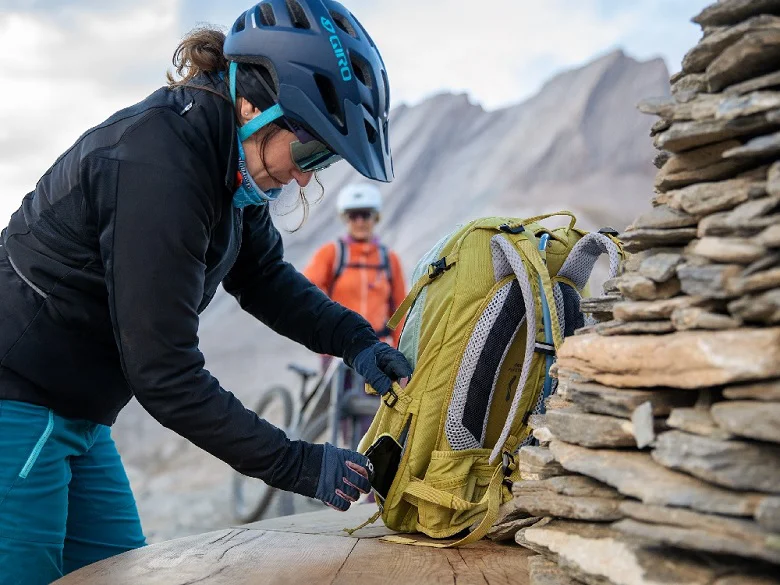 deuter Trans Alpine Fahrradrucksack Outdoor Imagebild - Person mit Fahrradbekleidung und Fahrradhelm bedient ein Außenfach eines gelben Rucksacks.