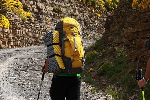 Trekkingrucksack richtig einstellen Person mit gelbem Rucksack wandert einen Kiesweg hinaut, am Rucksack ist eine gelbe Isomatte außen befestigt.