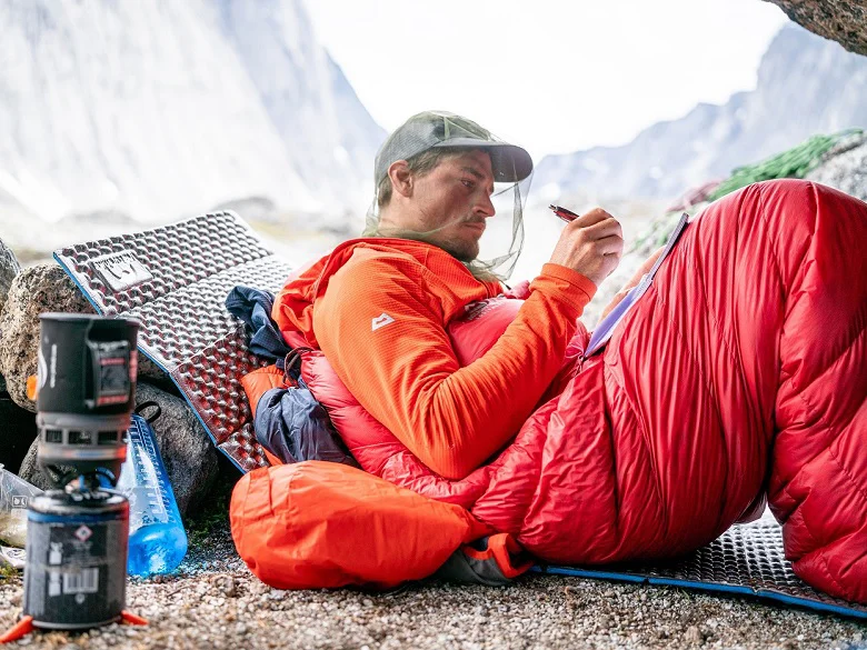 Mountain Equipment Glacier Outdoor Imagebild - Person liegt im freien auf einer Isomatte in einem roten Schlafsack. Sie trägt eine Kappe mit Moskitonetz, außerdem zu sehen eine Trinkflasche und ein Outdoorkocher mit Kartusche.