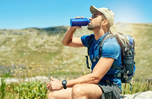 Person trinkt aus einer Flasche im Freien. Sie ist mit T-Shirt, Schildkappe und Shorts gekleidet und trägt einen Rucksack