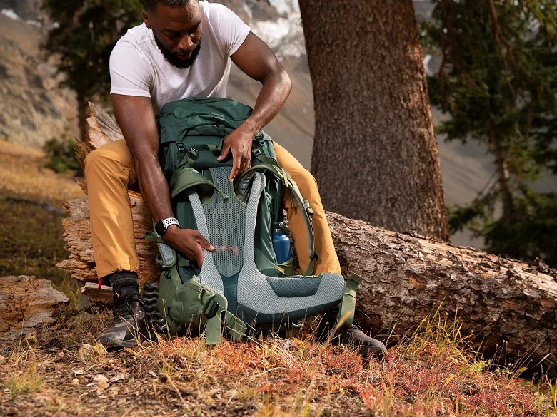 Trekkingrucksack richtig packen Person sitzt auf einem Baumstamm und hat einen grünen Rucksack. Sie verstellt den Rückenträger des Rucksacks.