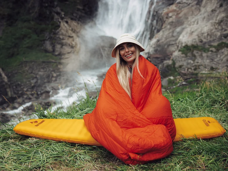 Outdoor Imagebild - Person sitzt auf einer gelben Isomatte eingewickelt in einem roten Schlafsack, im Hintergrund ein Wasserfall