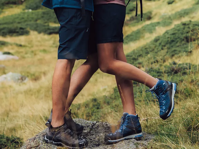 zwei Personen in kurzer Hose mit Wanderschuhen stehen auf einem Fels in den Bergen.