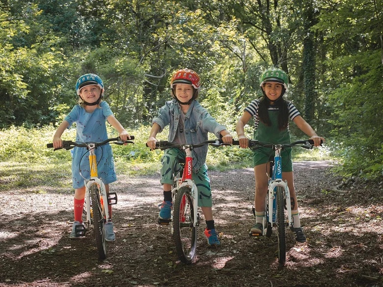 Woom Bikes - Kinderleicht Radfahren lernen Outdoor Imagebild - drei Kinder auf Woom Bikes in gelb, rot und blau