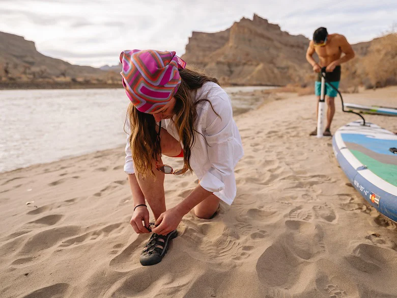 Outdoor Imagebild - zwei Personen an einem Strand, im Hintergrund Berge. Eine Person bindet sich die Schuhe im Vordergrund, eine zweite Person im Hintergrund pumpt ein Boot auf.