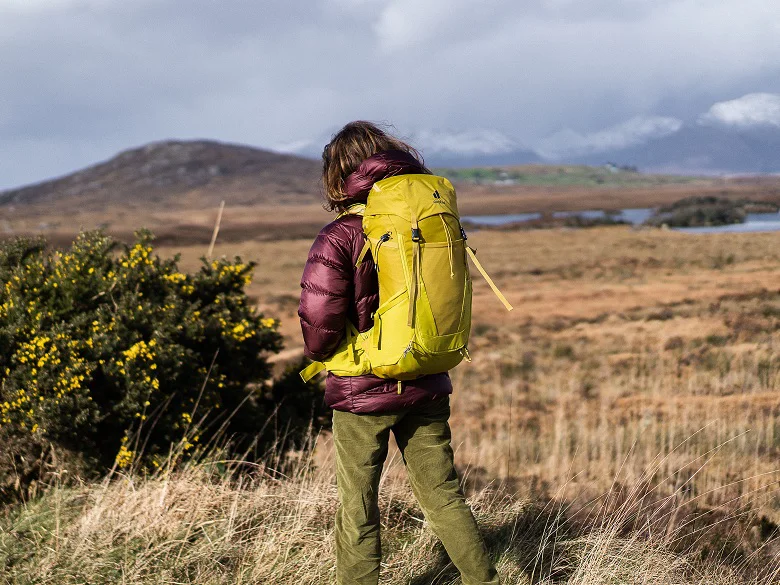 deuter Futura Rucksack Outdoor Imagebild - Person mit violetter Jacke und gelbem Wanderrucksack im freien in einer hügeligen Landschaft