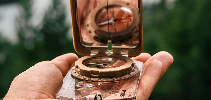 Outdoor Imagebild - Outdoor Kompass mit Spiegel liegt geöffnet auf einer Hand, im Hintergrund Berge und Natur